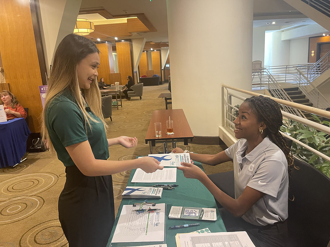 A Black woman with brown hair and professional clothing sitting down behind a table handing a brochure to a Pacific Islander woman with brown hair and green shirt who is standing on the opposite side of the table at an indoor event